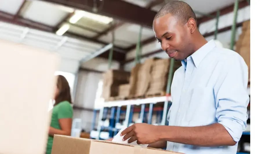 An African American man processing a packaged box in a warehouse, with another person in the background.